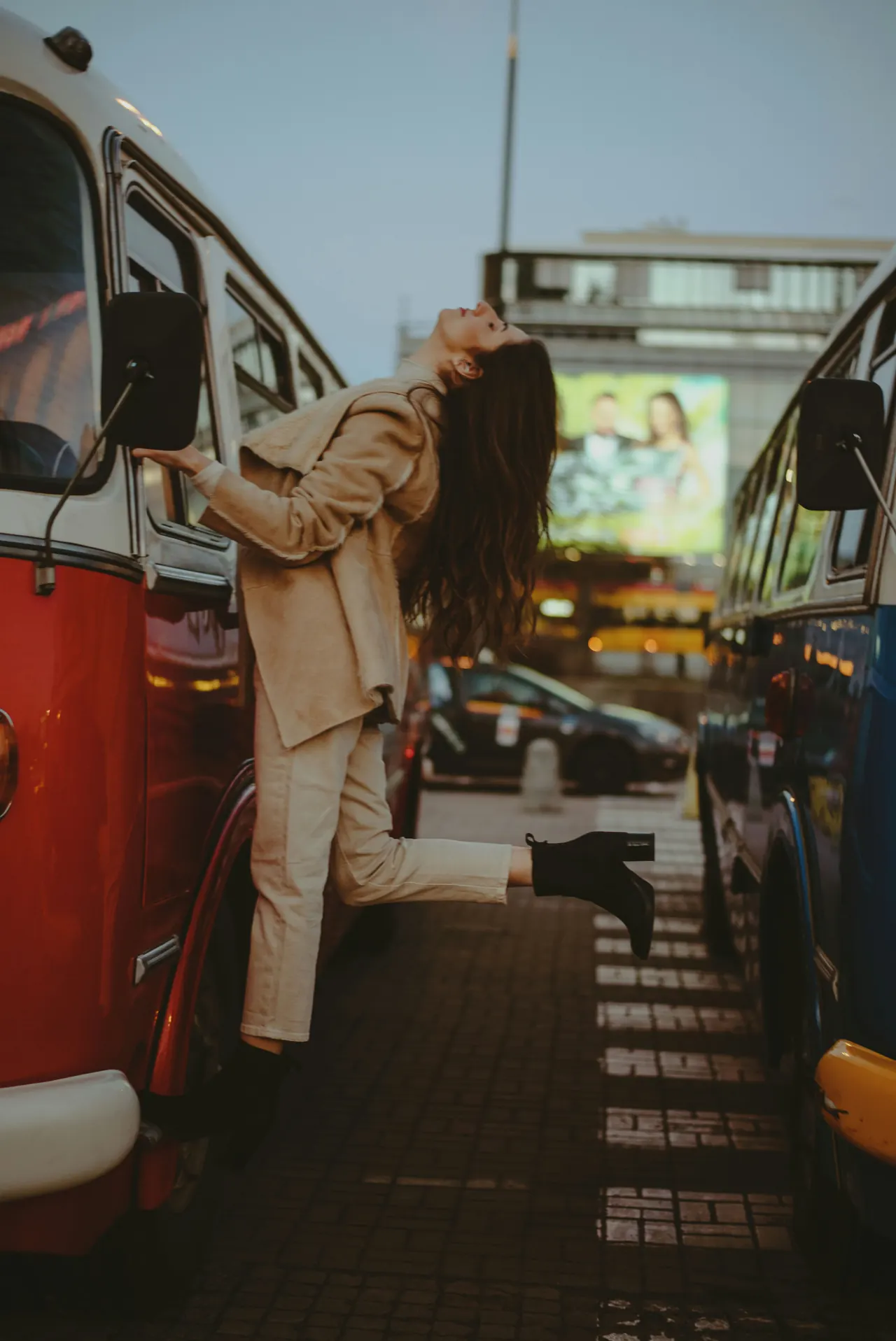 A woman leans back between two parked buses on a city street, her hair falling behind her.